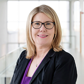 A woman wearing glasses and a black blazer stands in front of a window as she smiles at the camera.